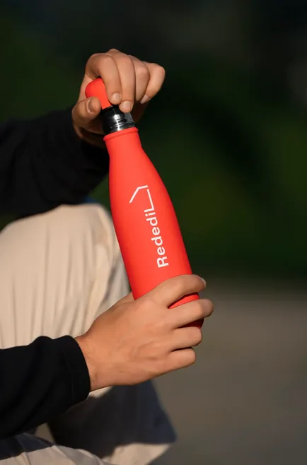 Borraccia rossa con il logo Rededil aperta a due mani, all’aperto